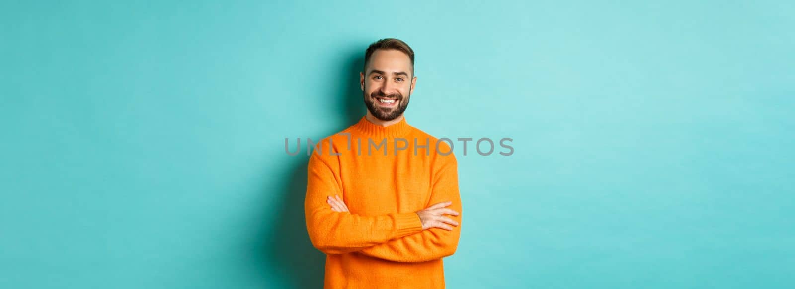 Handsome bearded male model in orange sweater, looking confident and smiling, cross arms on chest, standing over turquoise background.