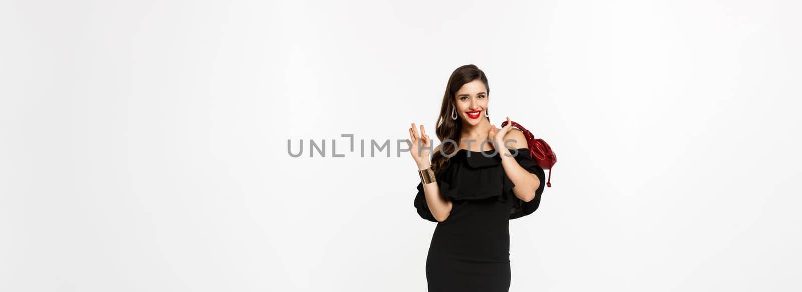 Beauty and fashion concept. Full length of confident and stylish young woman in glamour black dress, holding purse and walking forward, waving hand to say hello, white background.