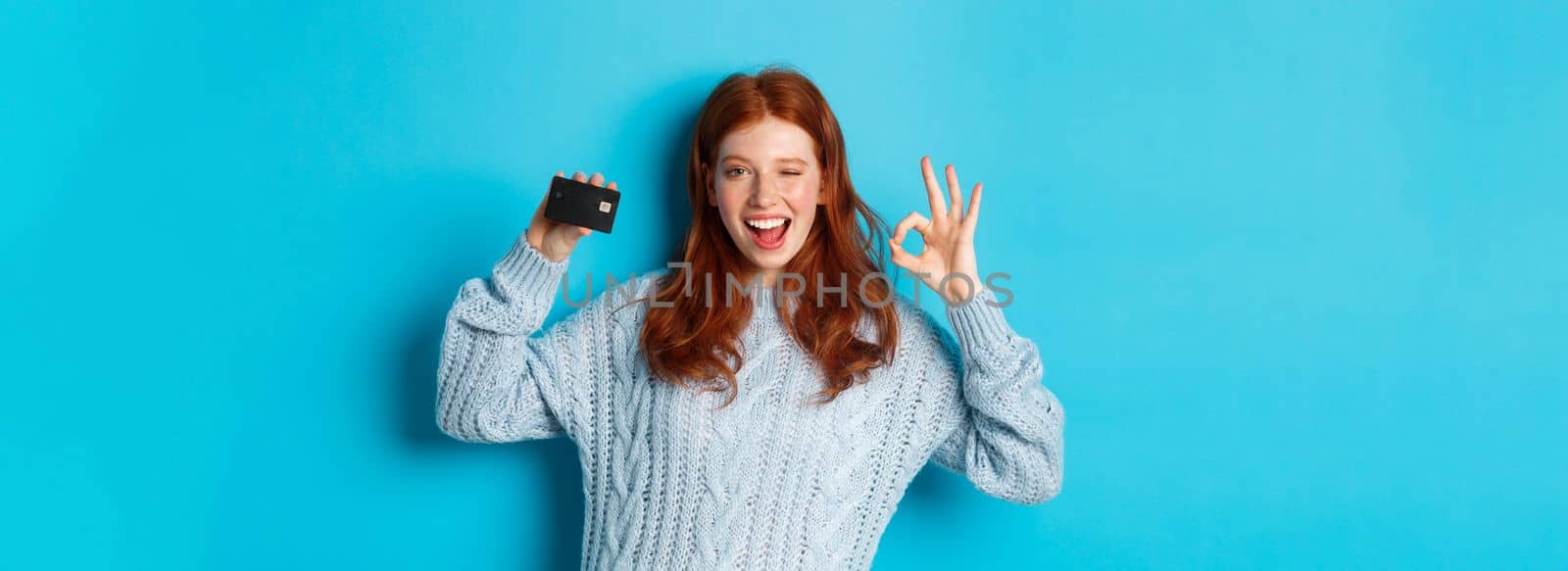 Happy redhead girl in sweater showing credit card and okay sign, recommending bank offer, standing over blue background.