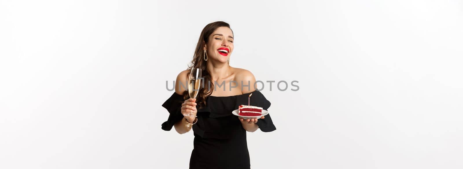Celebration and party concept. Fashionable woman holding birthday cake with candle and drinking champagne, laughing happy, standing over white background.