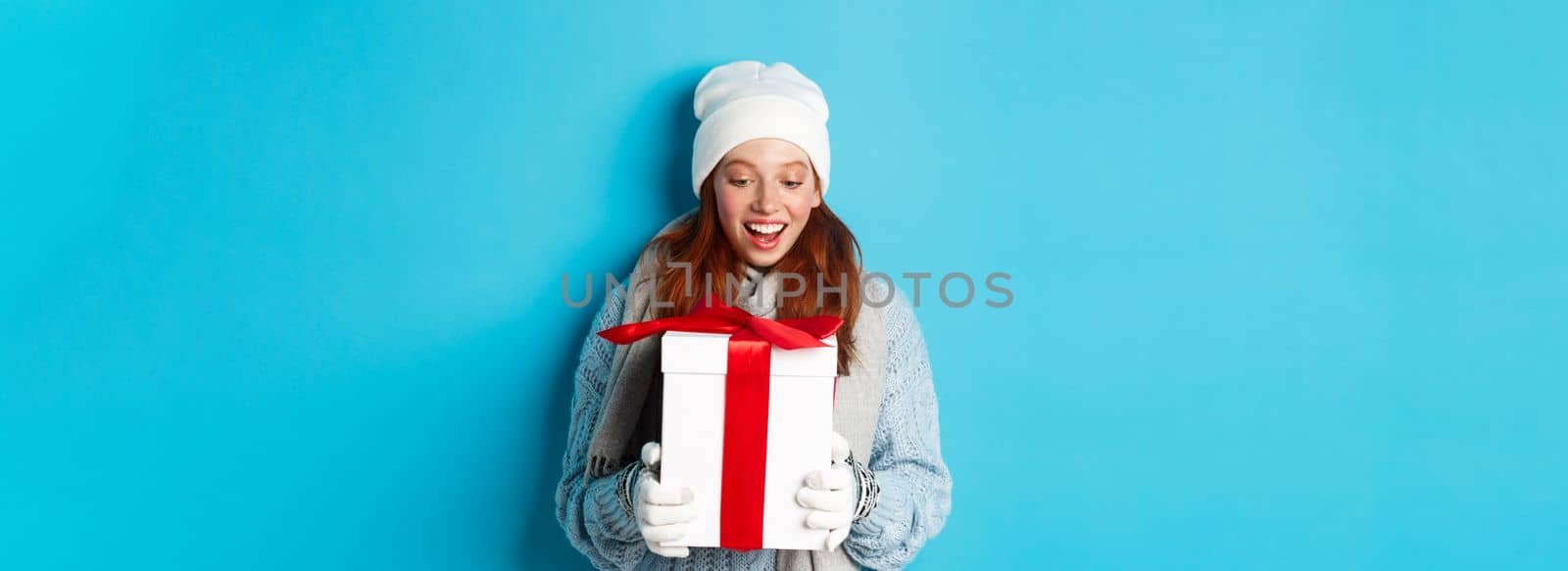 Winter holidays and Christmas eve concept. Surprised cute redhead girl in beanie and sweater receiving New Year gift, looking at present amazed, standing over blue background.