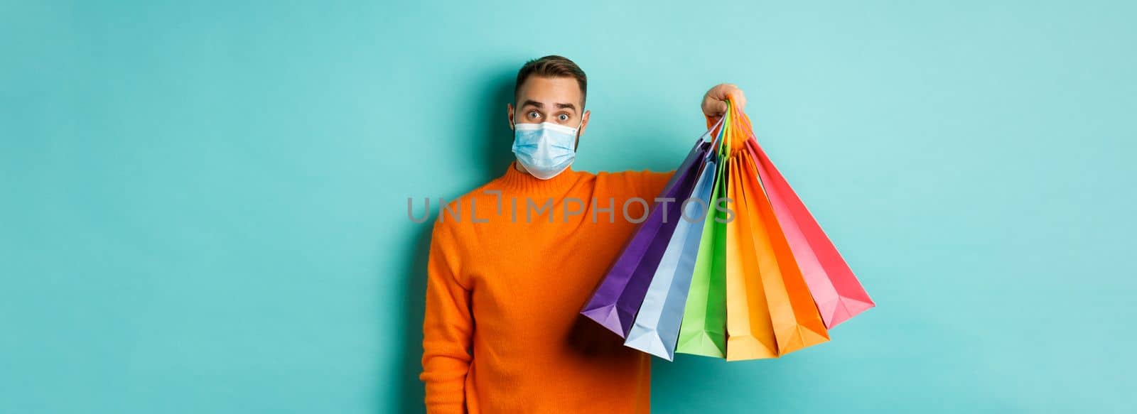 Covid-19, social distancing and lifestyle concept. Young man in face mask showing shopping bags, buying holiday gifts during pandemic, standing over blue background.