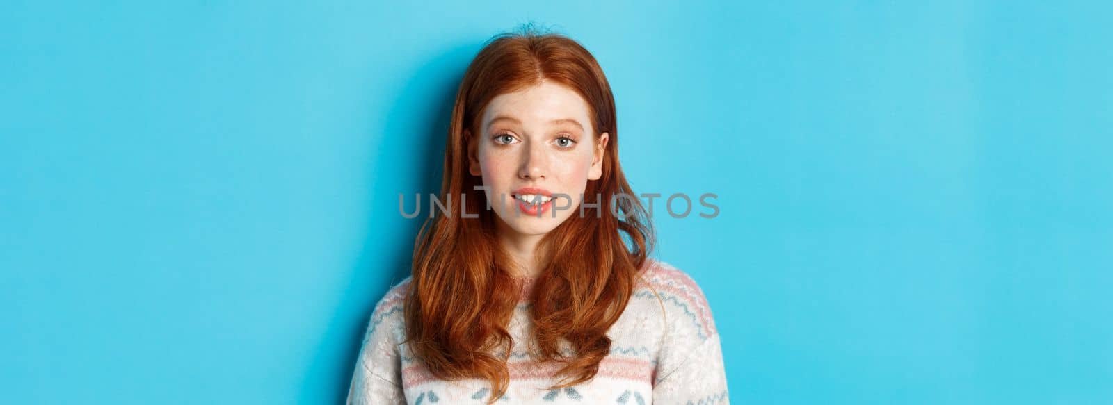Close-up of redhead girl looking intrigued at camera, hear something interesting, standing over blue background.
