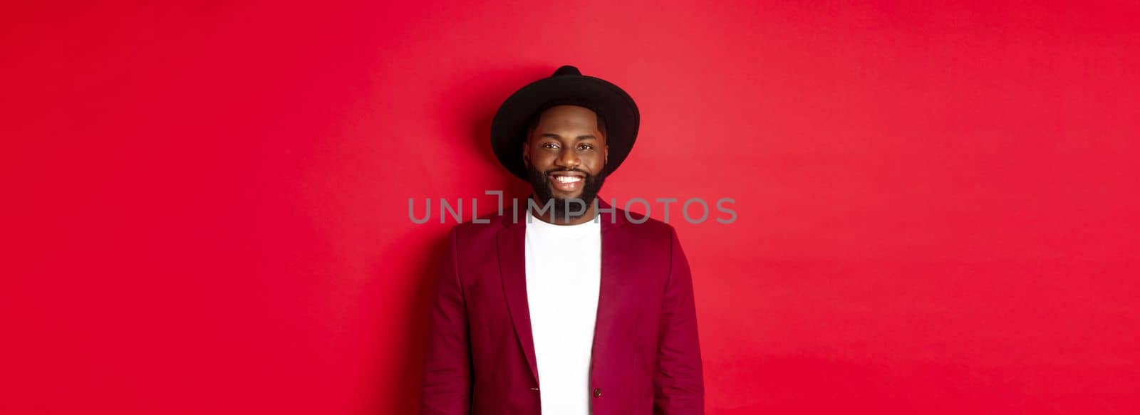 Fashion and party concept. Stylish african american man in blazer and hat, smiling confident at camera, celebrating new year, standing over red background.