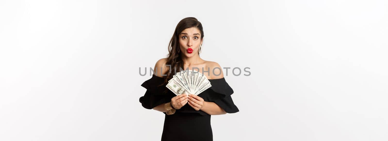 Fashion and shopping concept. Excited woman in black dress, with red lips, showing money dollars and looking amazed at camera, white background.