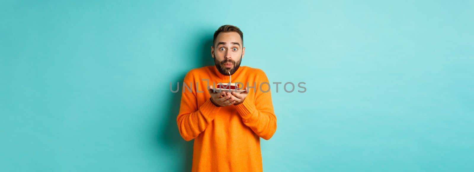 Handsome adult man celebrating birthday, blowing out candle on cake and making wish, standing against turquoise background.