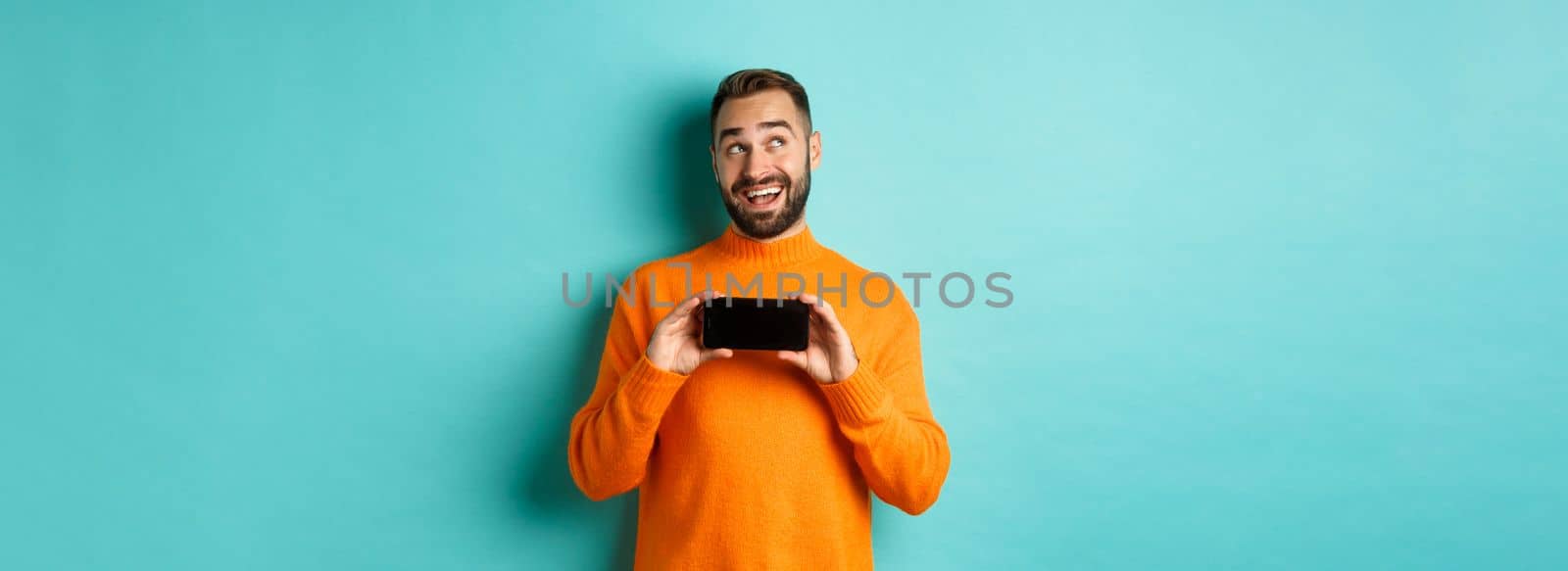 Online shopping. Young man thinking and showing smartphone screen, looking dreamy at upper left corner, standing over light blue background.