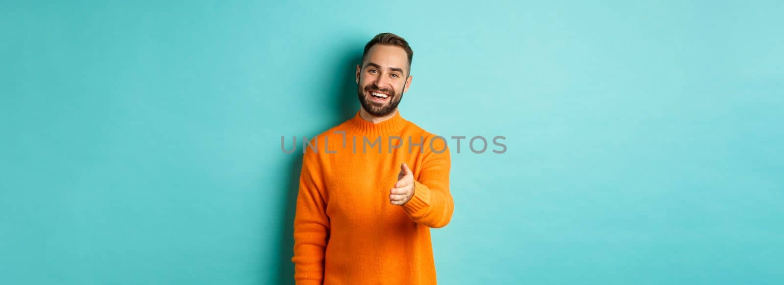 Friendly nice guy extend hand for handshake, greeting you and smiling, saying hello, standing over light blue background.