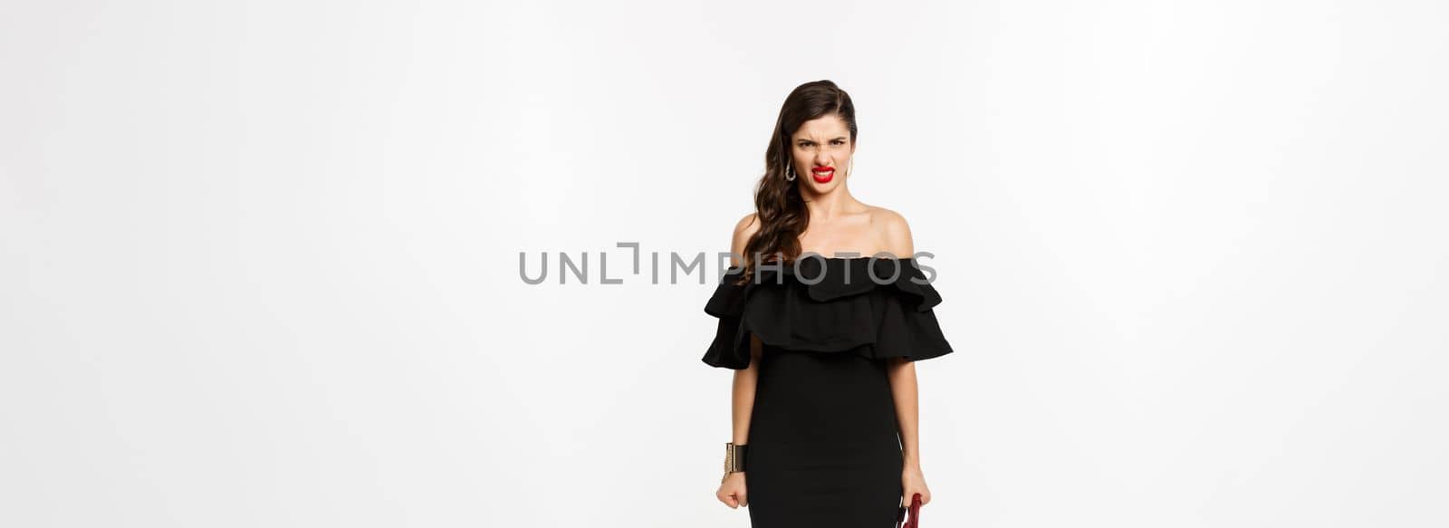 Beauty and fashion concept. Full length of angry woman in black party dress and high heels, express disdain and grimacing at camera, mad at person, white background.