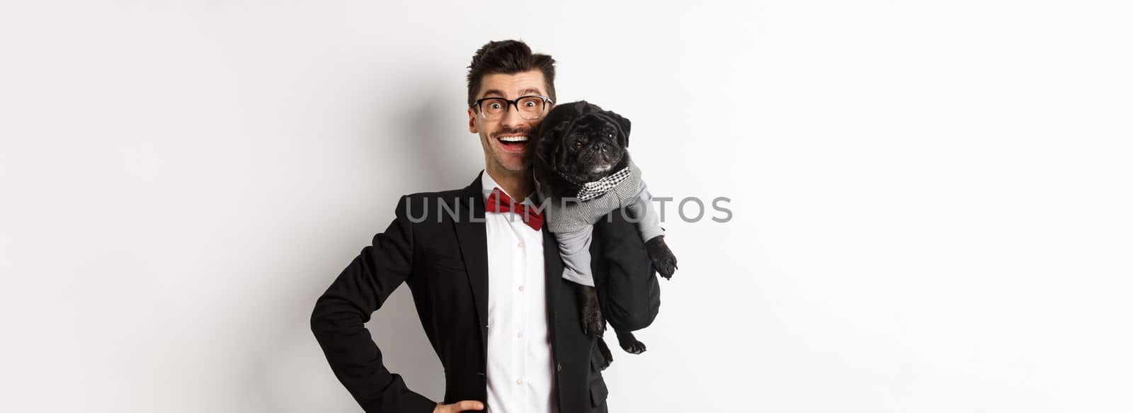 Handsome young man in suit and glasses holding cute black pug dog on shoulder, smiling happy at camera, wearing party outfits, white background.