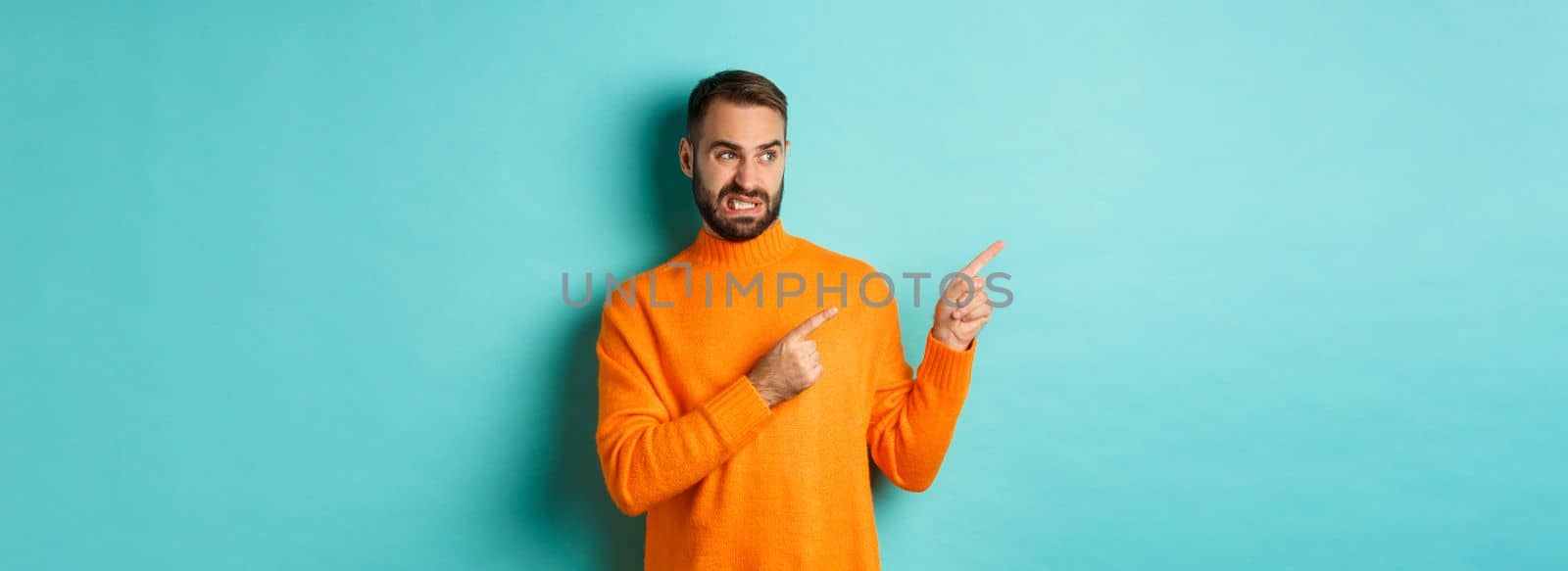 Displeased man cringe from something bad, pointing and looking at something disgusting or strange, left side copy space, standing against turquoise background.