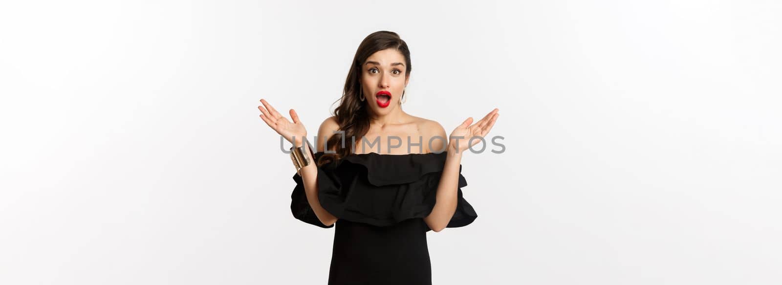 Beauty and fashion concept. Excited beautiful woman looking with amazement at surprise, reacting to good news, standing in black dress with makeup on, white background.