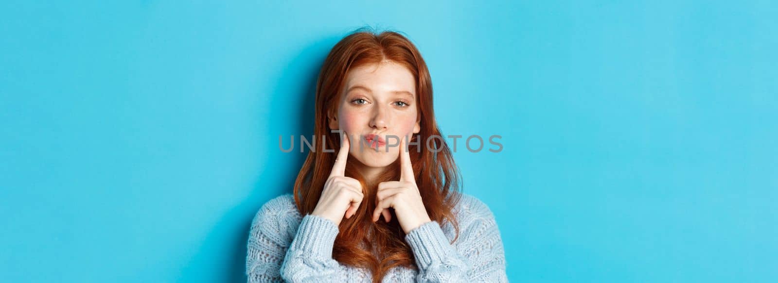 Close-up of cute redhead girl in sweater, pucker lips and pointing fingers at cheeks, poking dimples, standing over blue background.