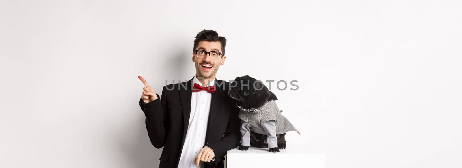 Animals, party and celebration concept. Happy and amazed young man and cute pug in costume standing over white background, dog owner pointing right.