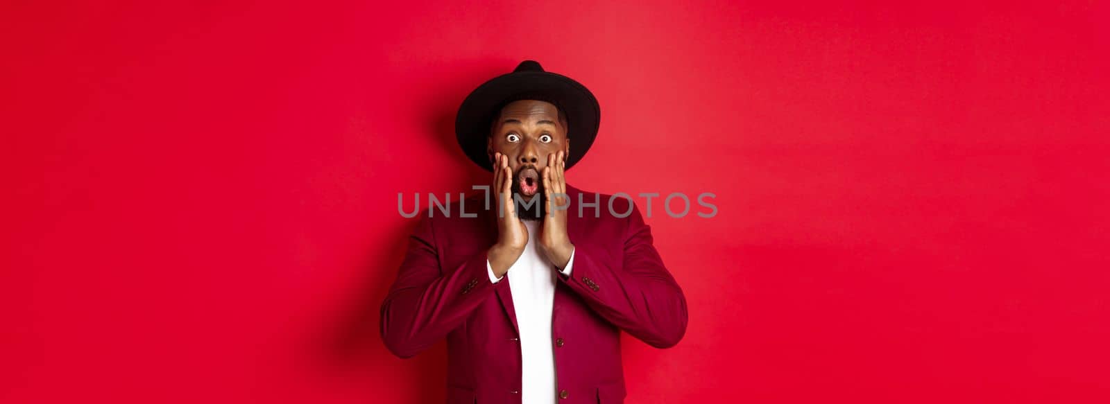 Fashion and party concept. Impressed Black man in classy outfit staring with complete disbelief at camera, gasping and looking surprised, standing over red background.