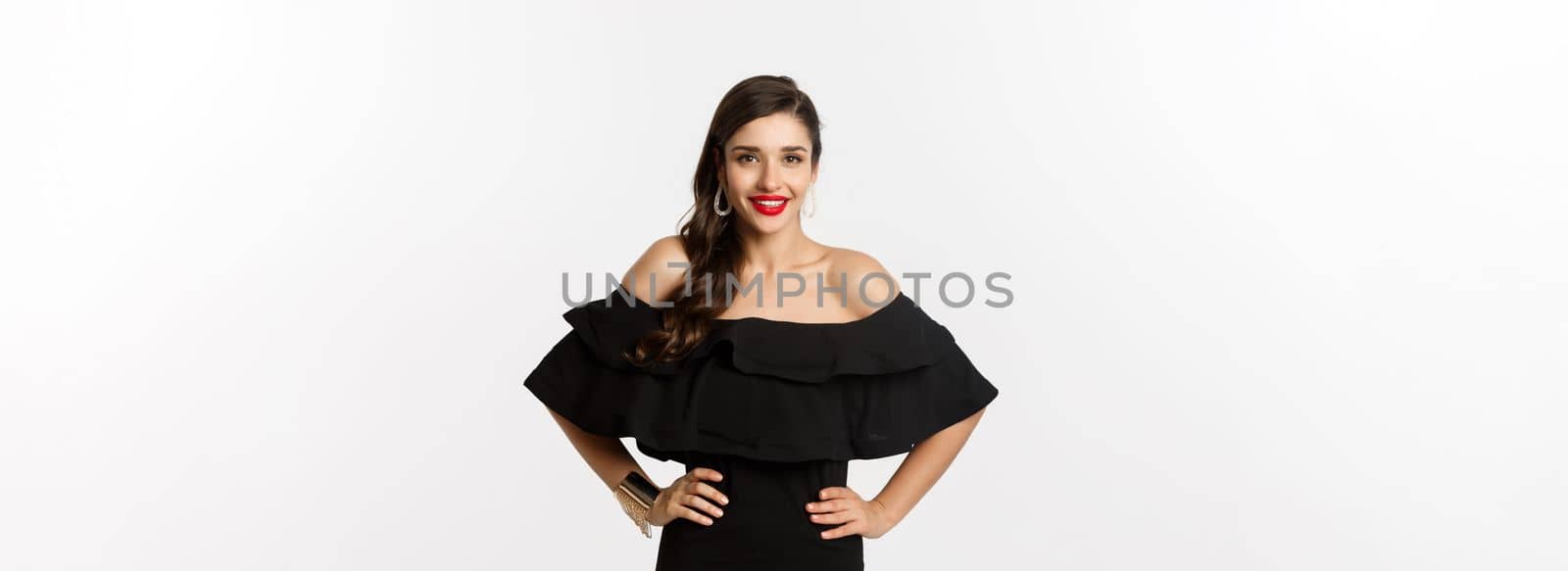 Waist-up shot of beautiful woman with red lipstick, wearing black dress and smiling pleased, standing over white background.