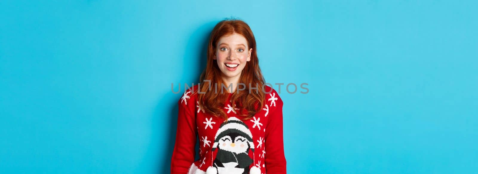 Winter holidays and Christmas Eve concept. Excited redhead girl in xmas sweater looking surprised at camera, standing against blue background by Benzoix