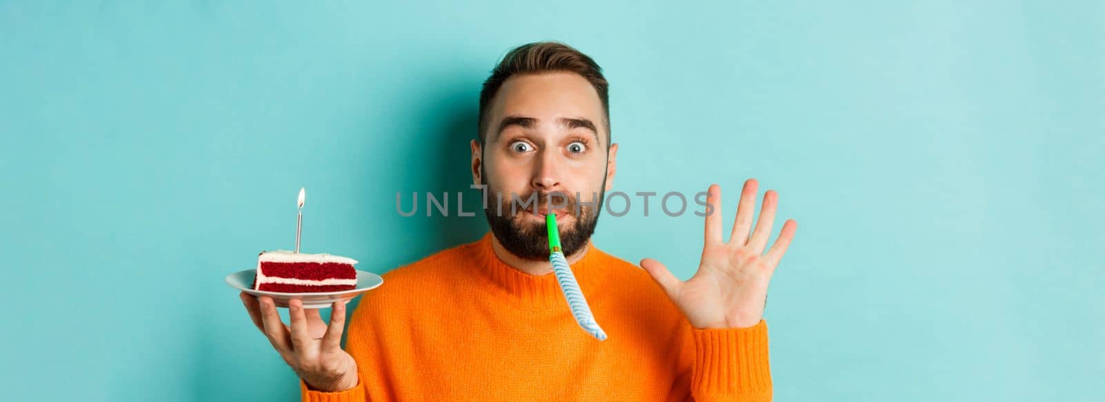 Close-up of funny adult man celebrating his birthday, holding bday cake with candle, blowing party wistle and rejoicing, standing over light blue background by Benzoix