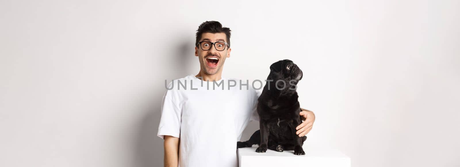 Image of amazed man staring at camera while his small dog looking left at copy space, standing over white background.