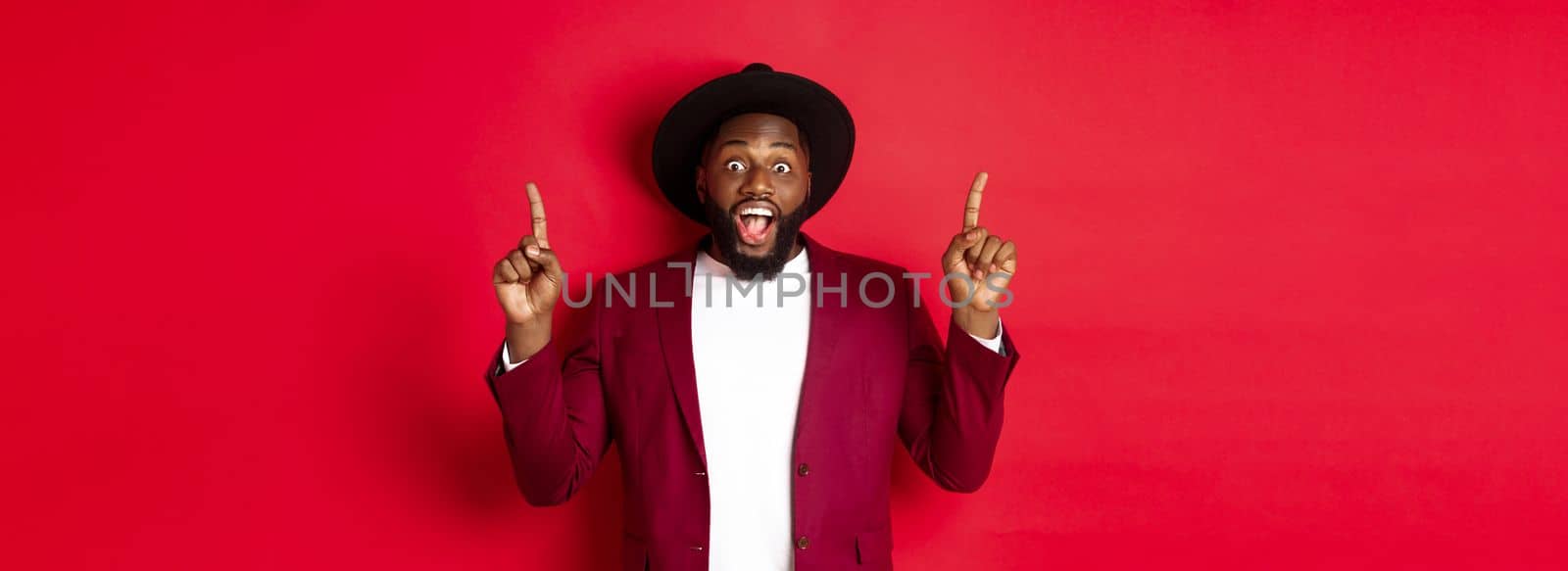 Winter holidays and shopping concept. Cheerful african american man in party outfit pointing fingers up, showing logo and smiling happy, red background.