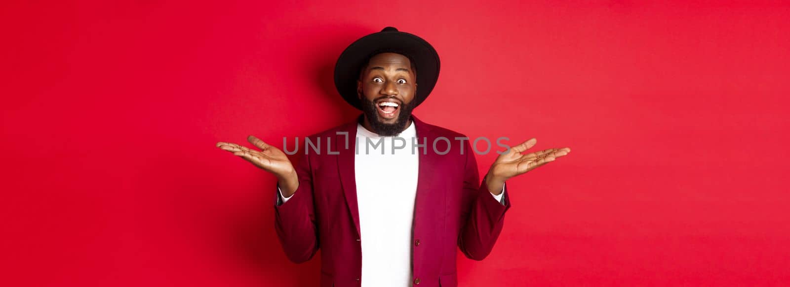 Christmas shopping and people concept. Handsome african american man smiling, spread hands sideways, showing promo offers on copy space, red background by Benzoix