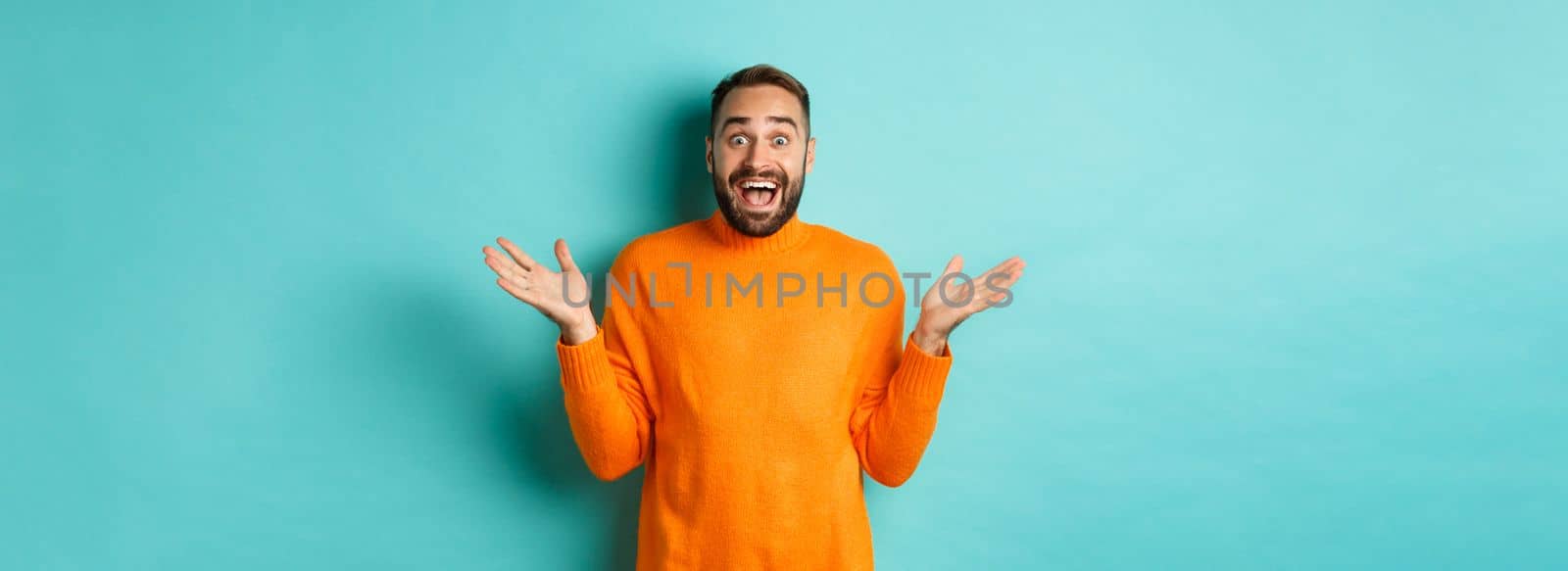 Surprised man raising hands up, looking at something amazing, standing over turquoise background in winter sweater by Benzoix