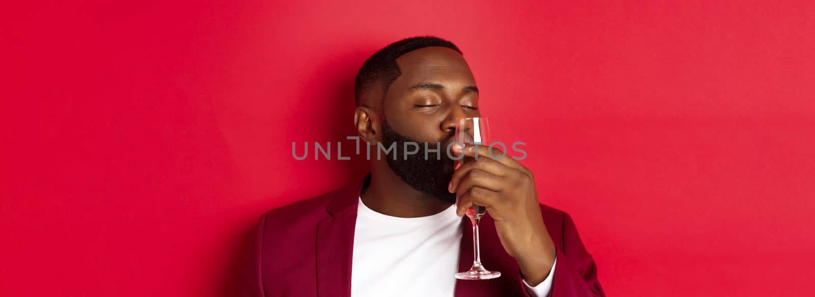 Drunk african american guy kissing glass with champagne, celebrating new year, enjoying party, standing over red background.