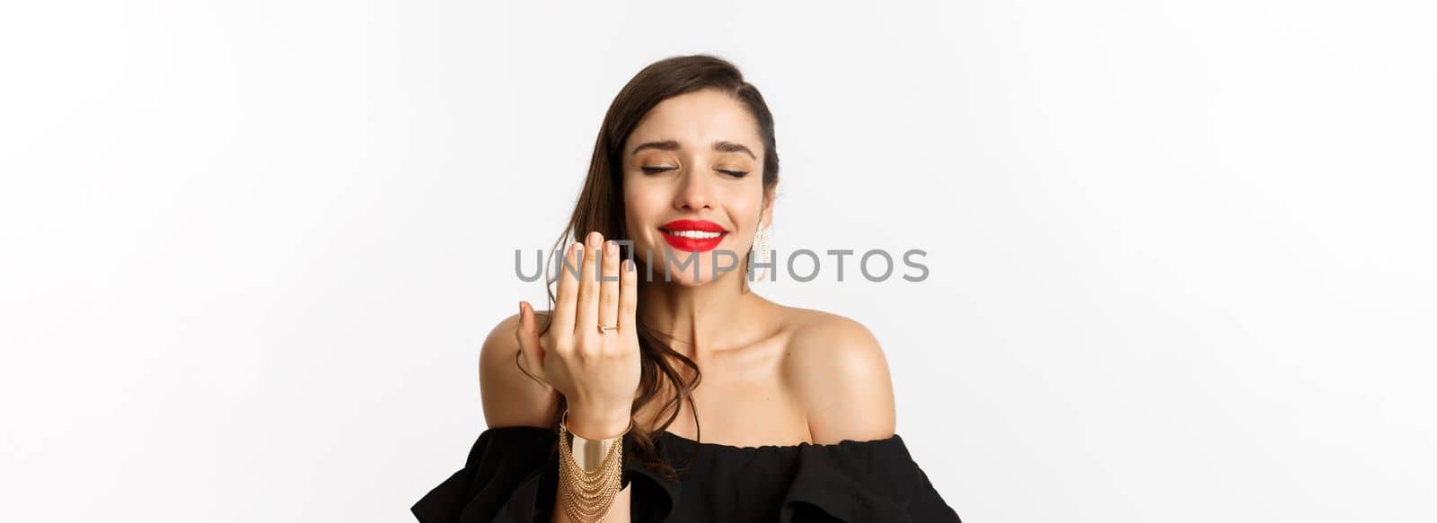 Happy young woman saying yes, become a bride, showing engagement ring on finger and smiling pleased, standing over white background by Benzoix