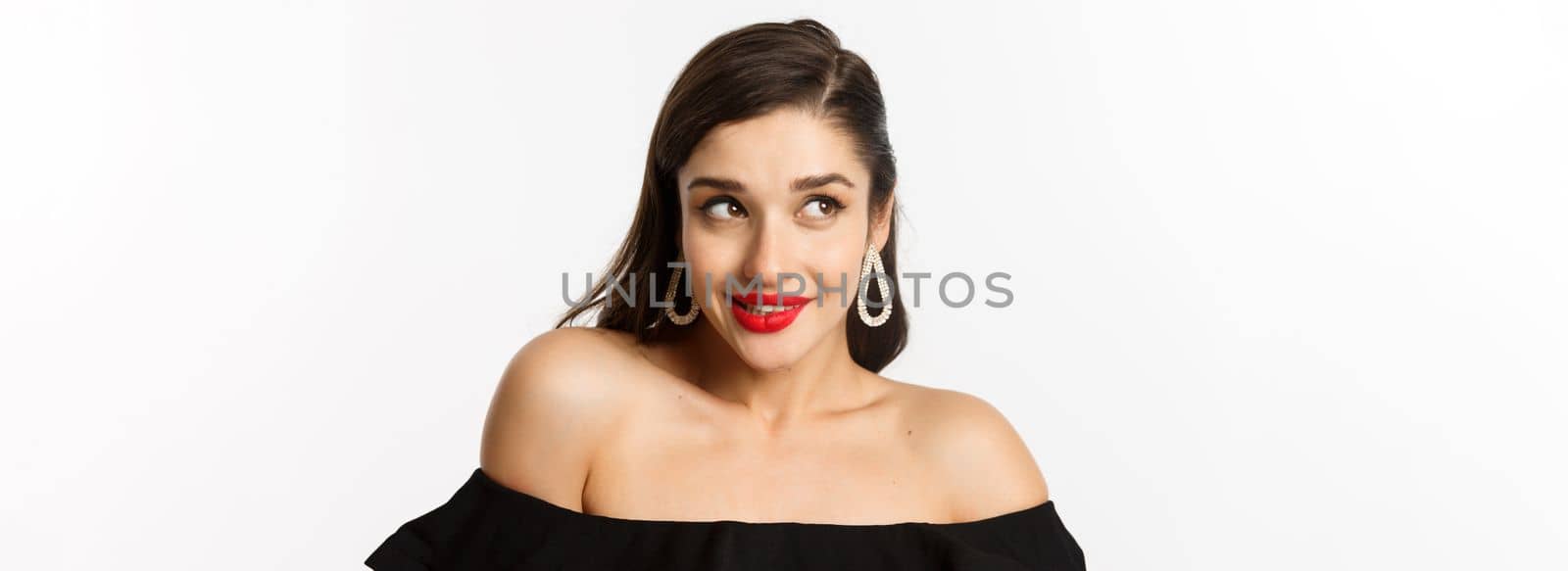 Fashion and beauty concept. Close-up of beautiful woman with red lips, makeup and black dress, showing her earrings and looking pleased, white background.