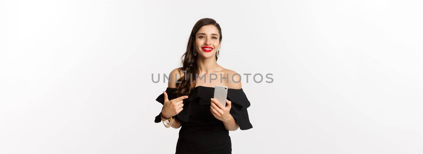 Online shopping concept. Stylish woman in black dress, wearing makeup, pointing finger at mobile phone with satisfied smile, standing over white background.