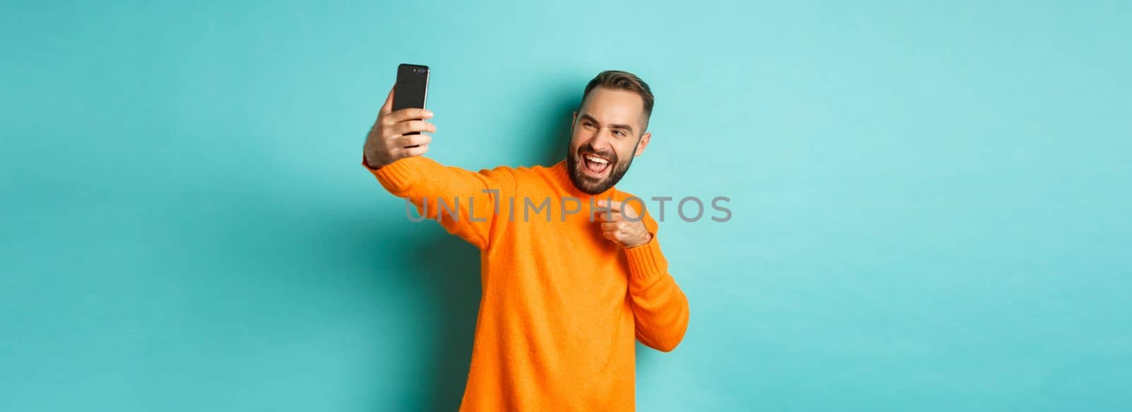 Handsome man video calling on mobile phone, taking selfie and pointing at smartphone camera, standing over turquoise background.