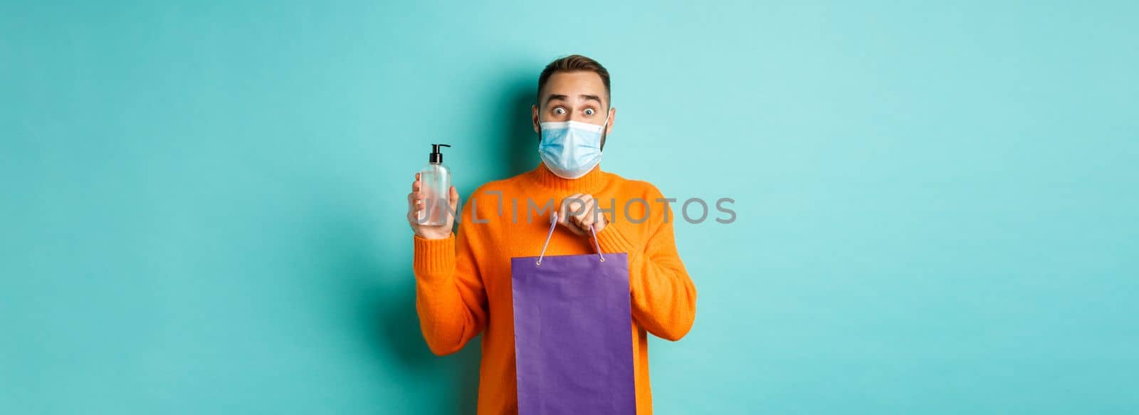 Coronavirus, pandemic and lifestyle concept. Man in face mask showing shopping bag and hand sanitizer, standing over turquoise background by Benzoix