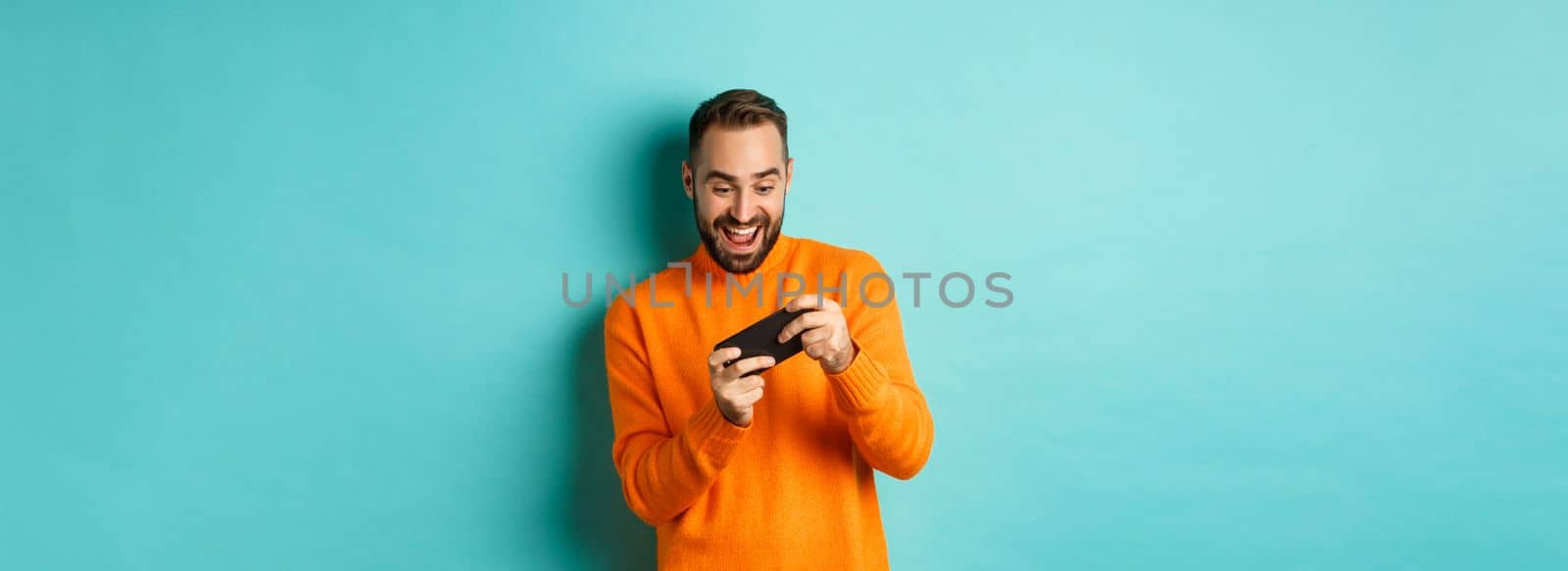Excited adult man playing on mobile phone, looking amazed at smartphone screen, standing over turquoise background by Benzoix
