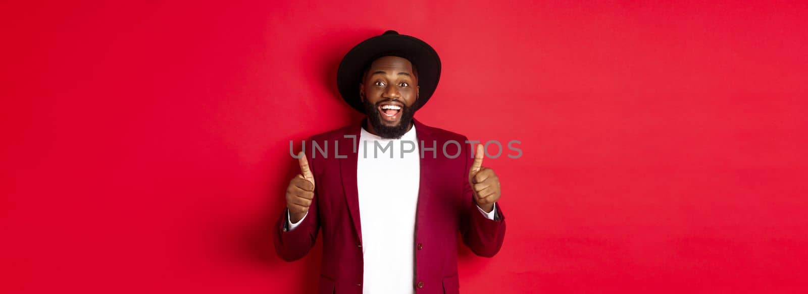 Christmas shopping and people concept. Handsome Black man smiling satisfied, showing thumbs-up, like and agree, approve something, standing against red background.