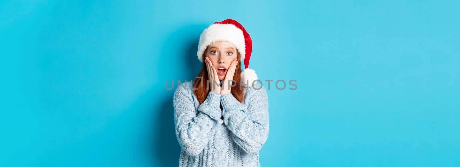 Winter holidays and Christmas Eve concept. Surprised redhead girl in santa hat, staring with disbelief at camera, open mouth amazed, standing over blue background by Benzoix
