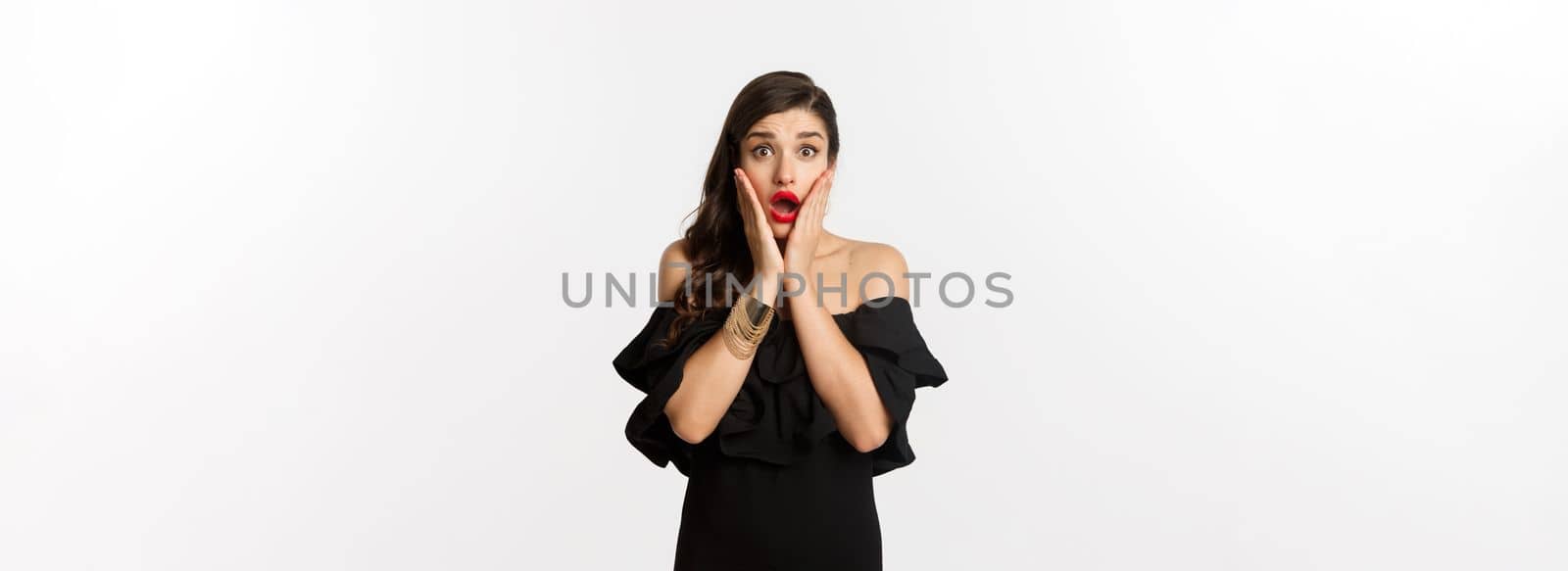 Beauty and fashion concept. Waist-up shot of surprised woman in black cocktail dress, looking amazed at camera, open mouth fascinated, standing over white background by Benzoix