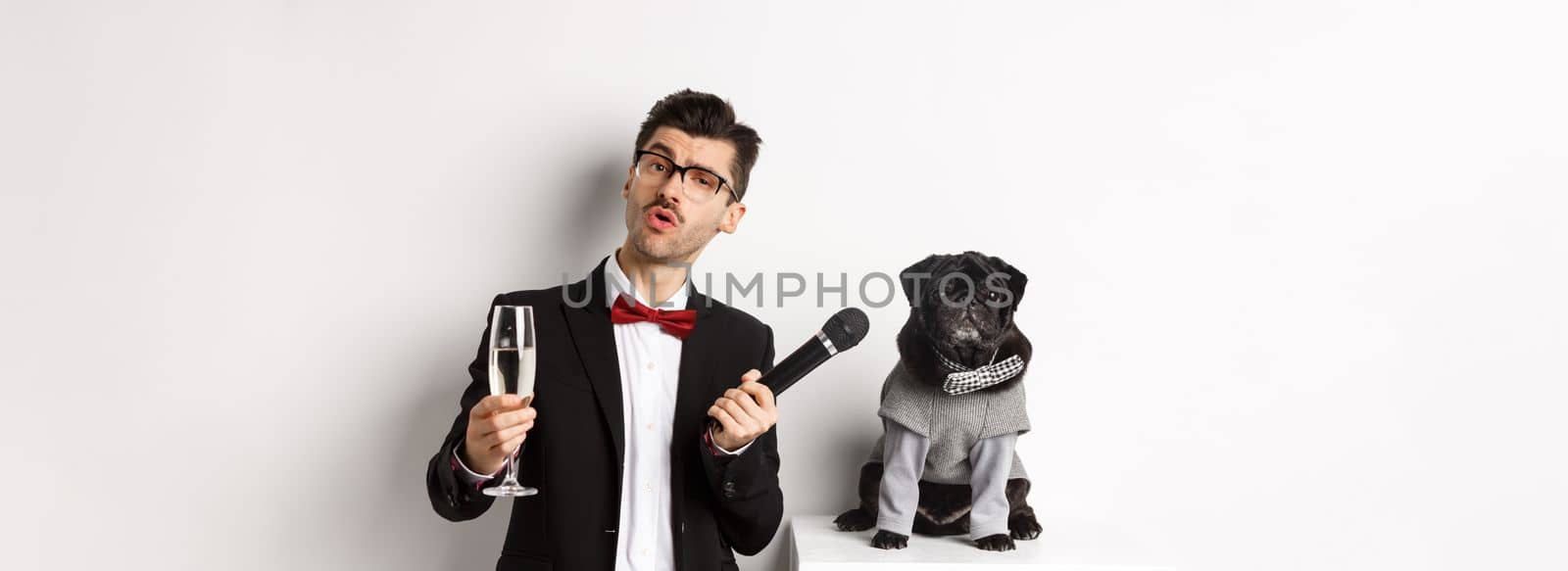 Handsome fancy man in glasses, raising glass of champagne and giving microphone to cute pug in party suit, celebrating and having fun, white background.