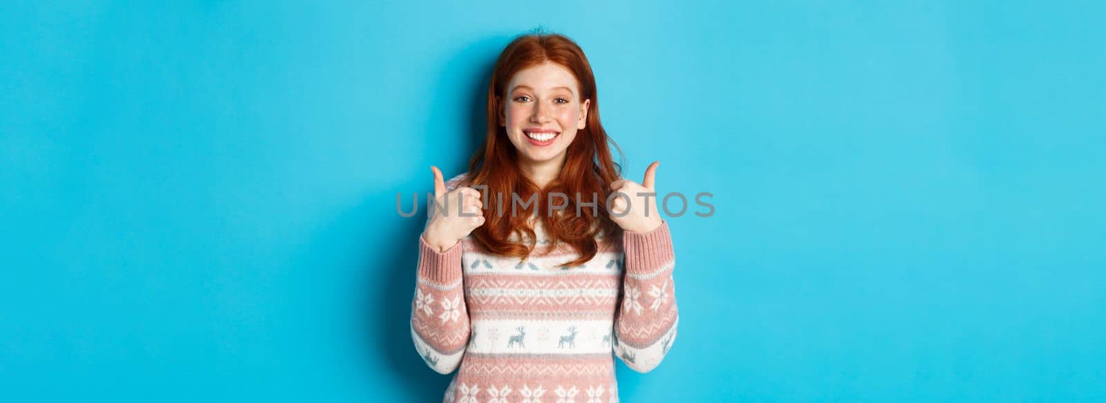 Cheerful redhead girl in winter sweater showing thumbs up, agree and like, showing her support and smiling, standing satisfied against blue background.