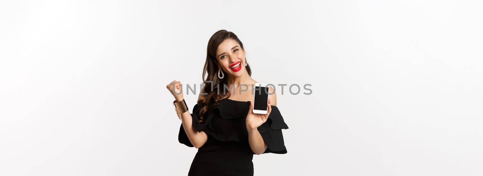 Online shopping concept. Satisfied pretty woman showing smartphone screen, making fist pump to rejoice, winning in internet, standing over white background by Benzoix