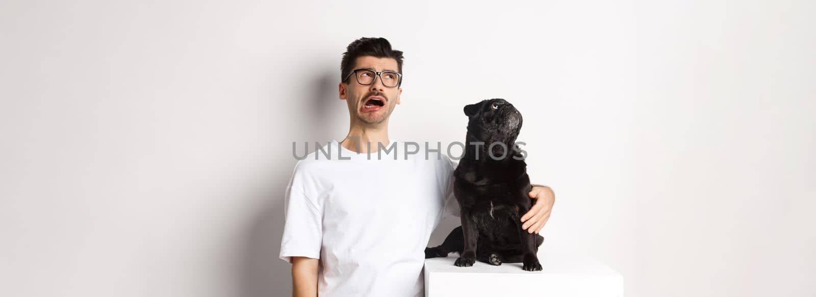 Whining man looking sad while pug staring intrigued at upper left corner, standing against white background.