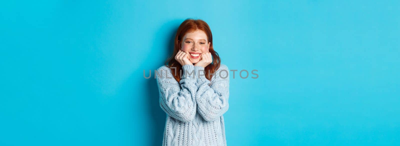 Lovely redhead teenage girl looking daydreaming, staring with admirationa sympathy at camera, standing against blue background by Benzoix