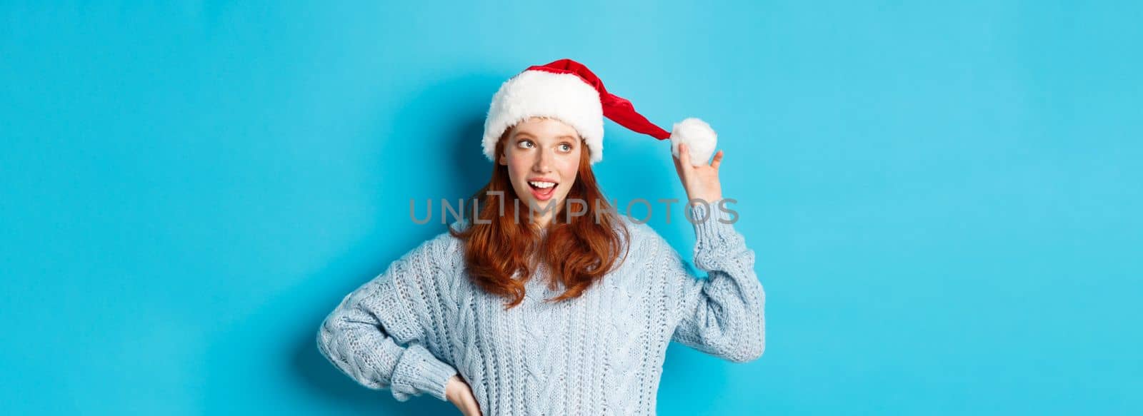 Winter holidays and Christmas Eve concept. Silly redhead girl with freckles, touching her santa hat and thinking, planning New Year celebration, standing over blue background.