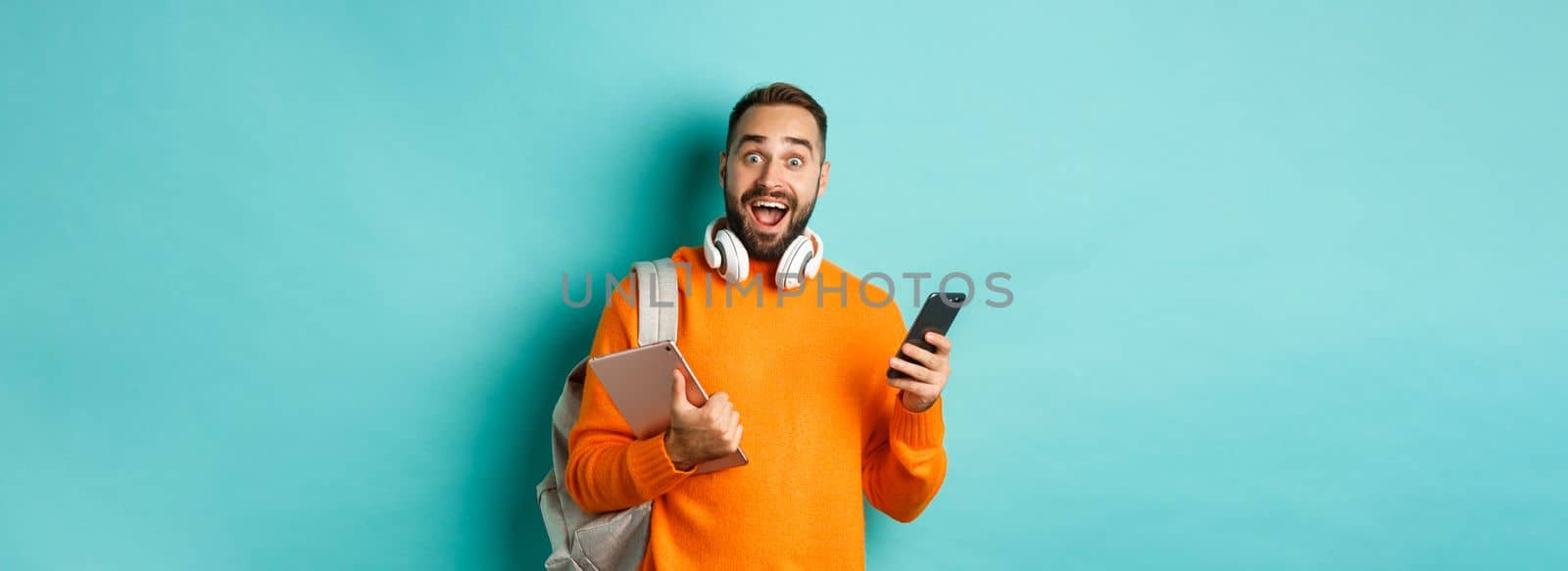 Handsome man student with headphones and backpack, holding digital tablet and smartphone, looking amazed at camera, standing against turquoise background.
