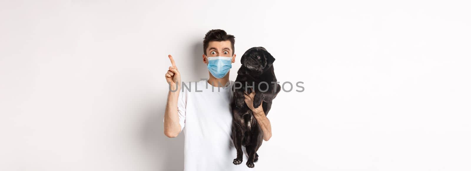 Covid-19, animals and quarantine concept. Young man in medical mask holding cute black pug, dog looking right and owner pointing at logo, white background by Benzoix