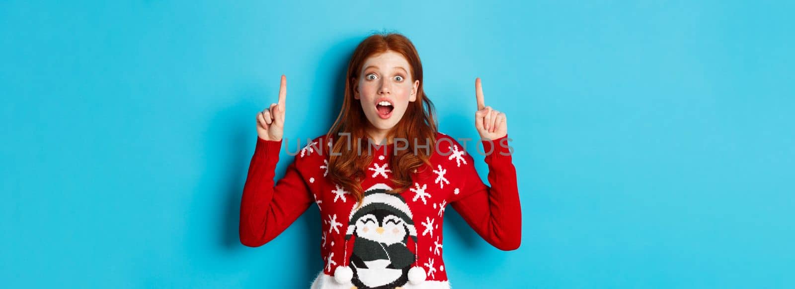 Winter holidays and celebration concept. Amazed redhead girl showing advertisement, pointing at christmas logo, standing excited over blue background by Benzoix