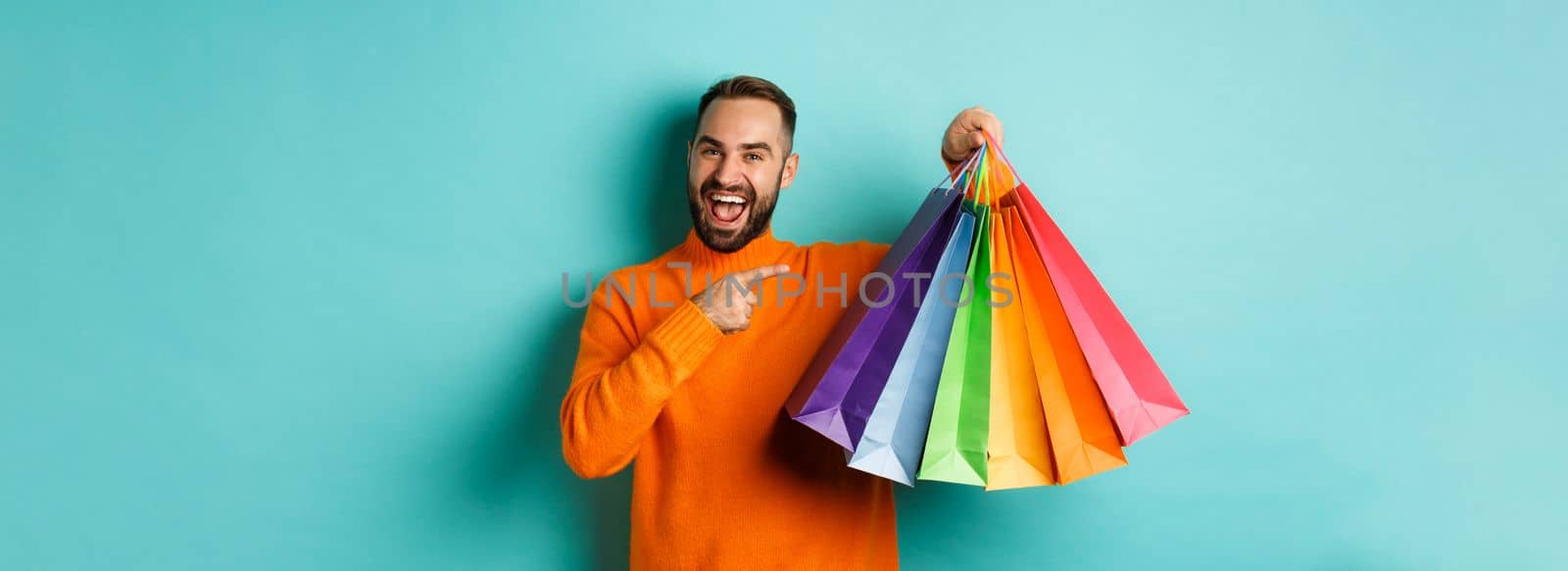 Happy handsome man holding shopping bags and smiling, pointing at purchased items and recommending store, standing over turquoise background.