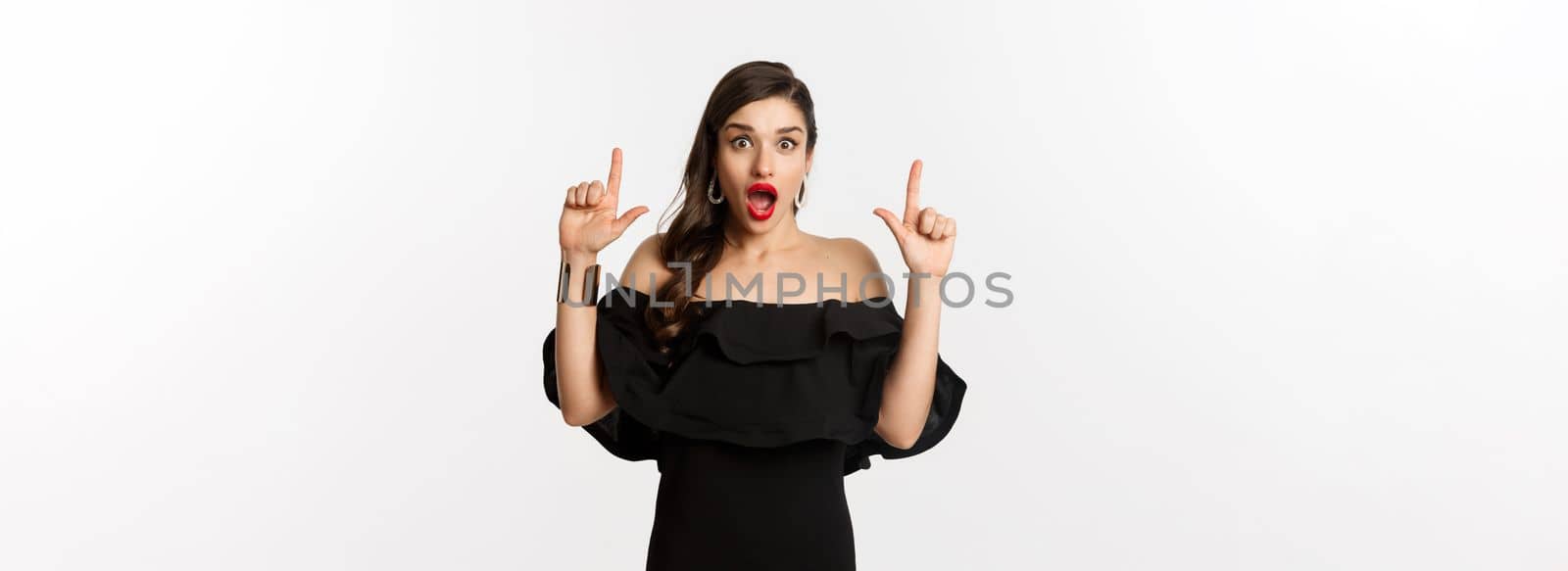 Fashion and beauty. Surprised woman in black dress pointing fingers up, showing banner, standing over white background.