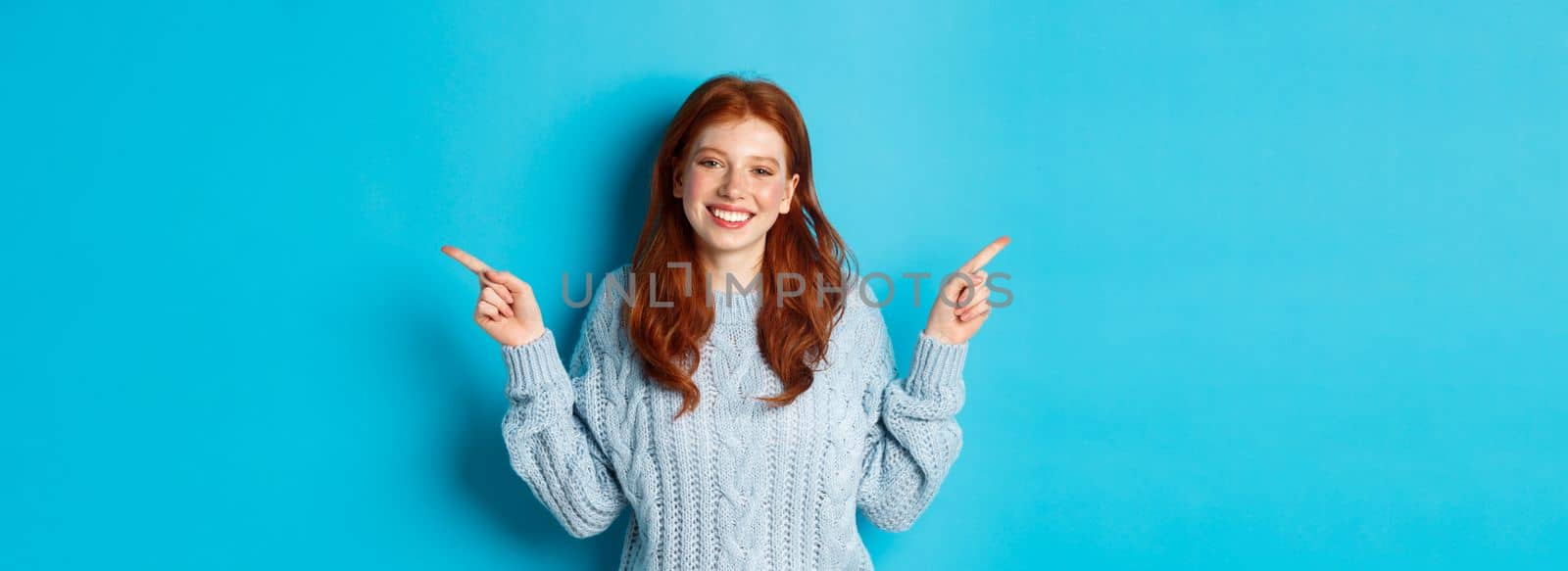 Winter holidays and people concept. Cute teen girl with red hair, smiling and pointing fingers sideways, showing advertisements, standing over blue background.