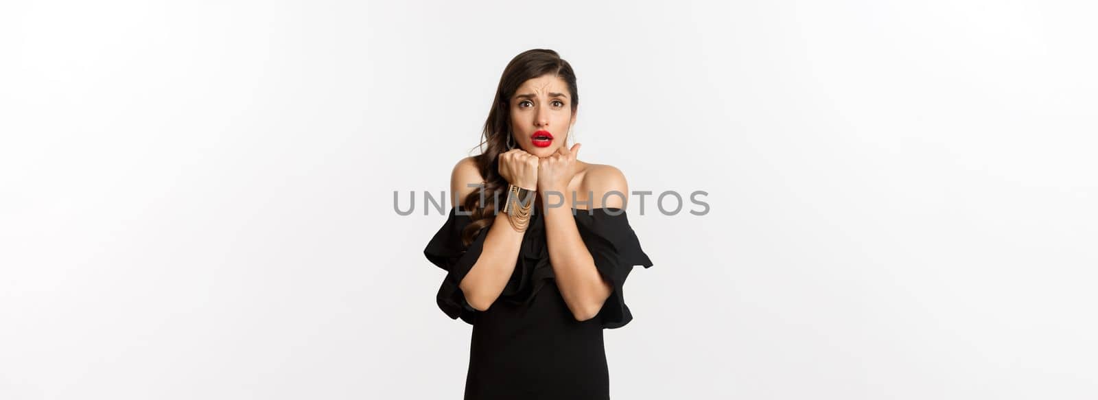 Fashion and beauty concept. Scared timid woman grimacing, looking concerned and worried at camera, standing over white background in black dress.