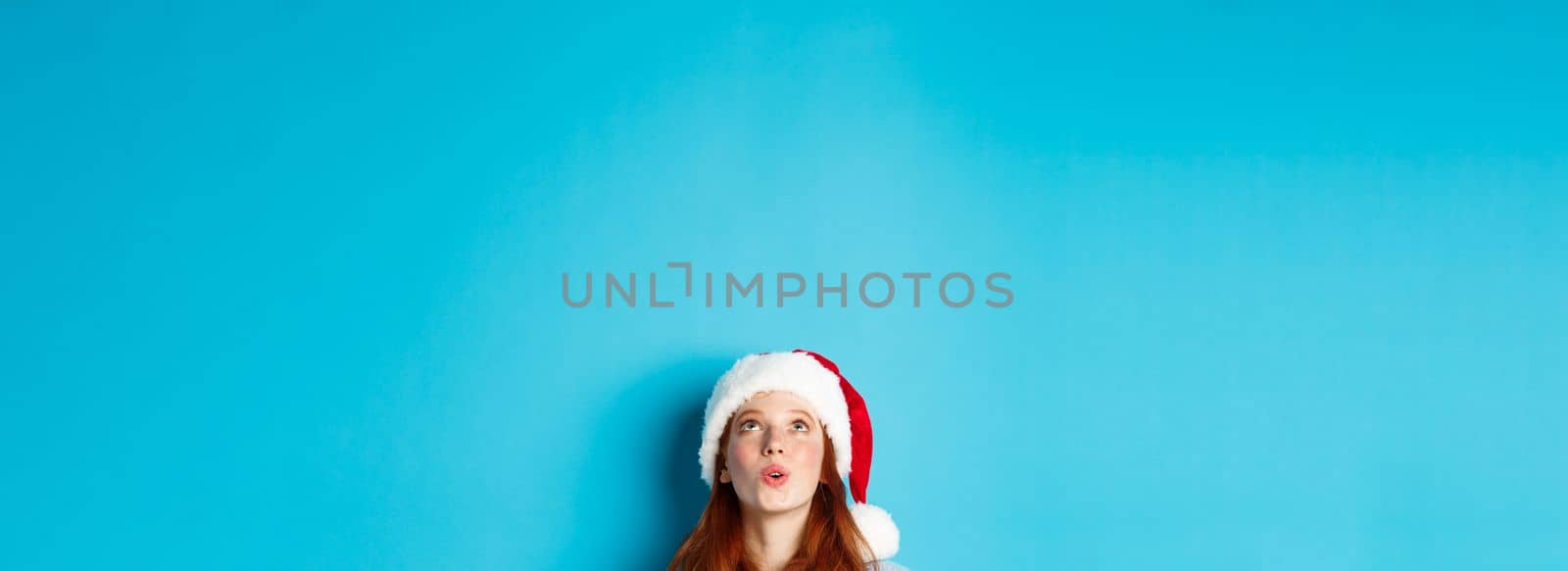Winter holidays and Christmas eve concept. Head of pretty redhead girl in santa hat, appear from bottom and looking up at logo impressed, seeing promo offer, blue background.